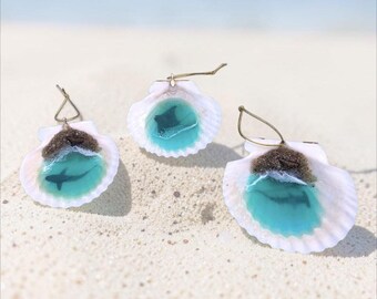 May include: Three seashell earrings featuring a resin-filled interior that mimics a miniature ocean scene. Each shell contains a turquoise blue water effect, a sandy beach area, and a small silhouette of a shark or a stingray.