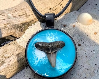 May include: A circular pendant necklace featuring a dark, fossilized shark tooth embedded in a bright turquoise resin background with white swirling patterns, suspended from a simple black cord.