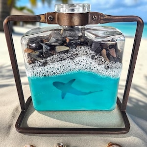 May include: A decorative glass bottle with a shark design, filled with blue liquid and small stones, set in a bronze-colored metal frame. The bottle has a clear glass stopper. Seashells are scattered on the sand in front of the bottle.