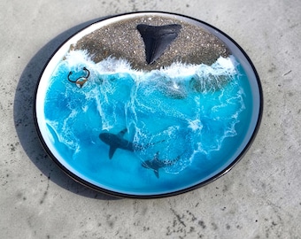 May include: A round decorative plate with a black rim, featuring a seascape design. The plate depicts a sandy beach, ocean waves, and two sharks swimming in the blue water. A large black shark tooth is embedded in the sand.