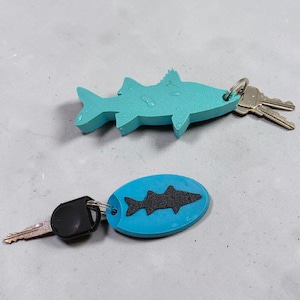 May include: A teal fish-shaped keychain with a metal ring holding two silver keys, and a blue oval keychain featuring a black textured fish silhouette attached to a black plastic key head with a metal ring.