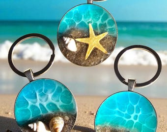 May include: Three round keychains with beach-themed designs. Each keychain features a silver-colored ring and a pendant with a sandy base, seashells, and a starfish or a shark tooth, all encased in clear resin with a blue ocean design.