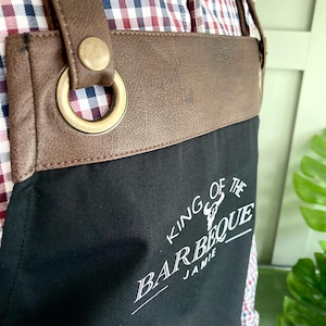 May include: Black canvas apron with brown leather straps and a brass buckle. The apron has the text "King of the Barbeque Jamie" printed in white.