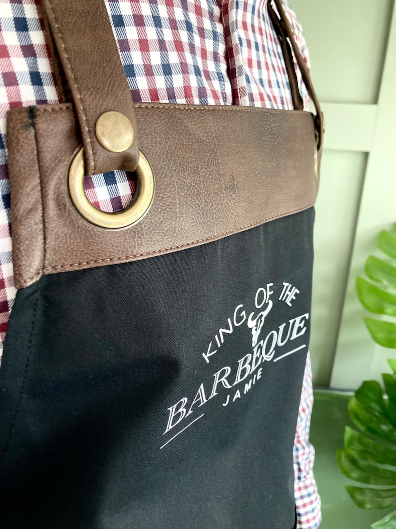 May include: Black canvas apron with brown leather straps and a brass buckle. The apron has the text "King of the Barbeque Jamie" printed in white.