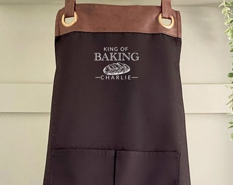 Puede incluir: Delantal negro con ribete de cuero marrón y tirantes ajustables. El delantal tiene un bolsillo grande y dice "King of Baking - Charlie" en texto blanco.