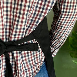 May include: A black apron with adjustable straps tied in the back. The apron is worn over a red and blue plaid shirt and blue jeans.