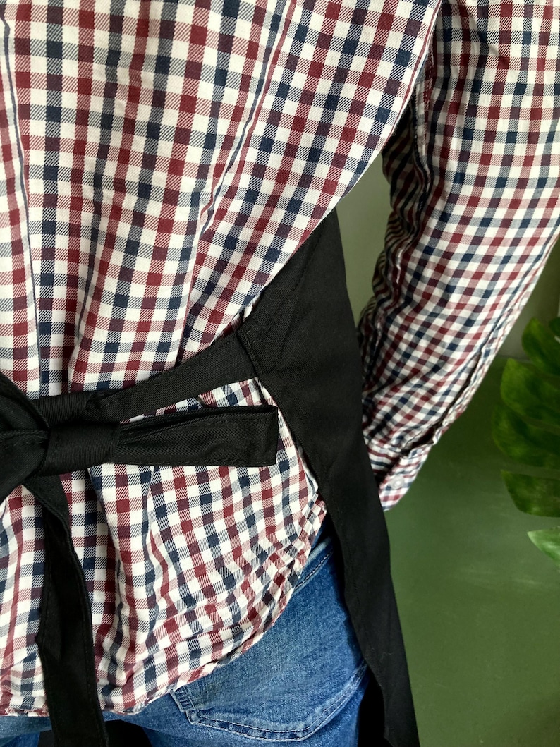 May include: A black apron with adjustable straps tied in the back. The apron is worn over a red and blue plaid shirt and blue jeans.