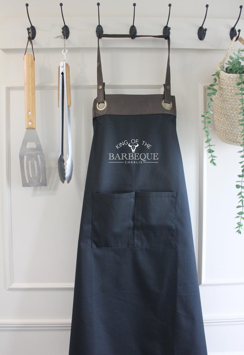 May include: Black apron with brown leather straps and two front pockets. The apron has the text "KING OF THE BARBEQUE - CHARLIE" printed in white. A spatula and tongs hang on the wall behind the apron.