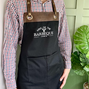 May include: Black apron with brown leather straps and a pocket. The apron has a white embroidered logo that says "King of the Barbeque Jamie".