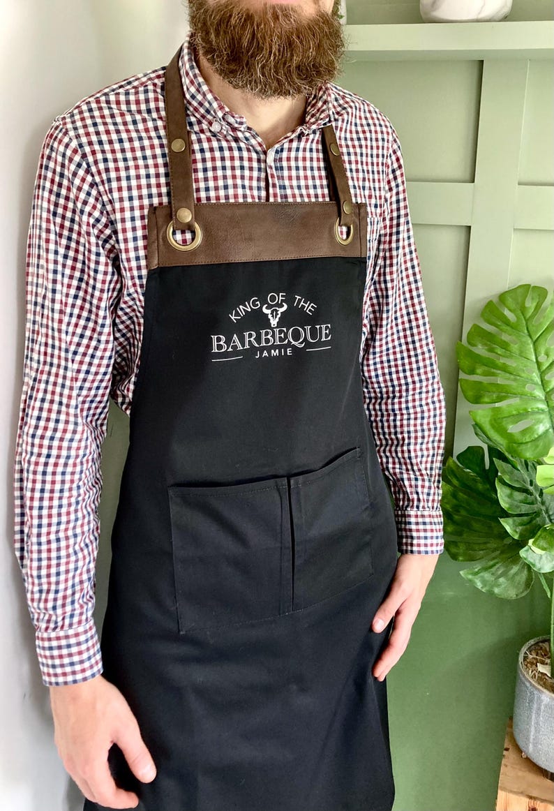May include: Black apron with brown leather straps and a pocket. The apron has a white embroidered logo that says "King of the Barbeque Jamie".