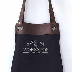 Puede incluir: Delantal de lona negro con ribetes de cuero marrón y correas ajustables. El delantal tiene un logotipo blanco que dice "King of the Workshop Charlie".