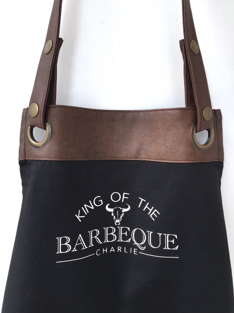 May include: Black canvas apron with brown leather straps and a white graphic that says "King of the Barbeque Charlie".