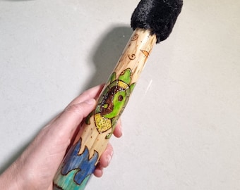 May include: A wooden stick with a black fur pom-pom on one end. The stick is decorated with a green and yellow turtle with a yin-yang symbol on its shell. The turtle is surrounded by blue and green waves.