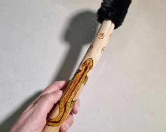 May include: A hand-painted wooden wand featuring a lizard design in yellow, orange, and green. The wand has a black fur handle and a carved end with a star design. The wand is approximately 12 inches long.