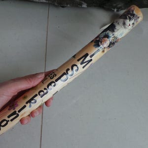 May include: A wooden rolling pin with a decorative design featuring a cat, flowers, and the words "Souvenirs". The rolling pin is light brown with black lettering and colourful floral accents.