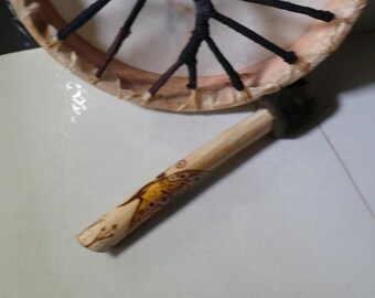 May include: A wooden frame drum with a natural leather head and a woven design. The drum is accompanied by a wooden beater with a carved butterfly design.