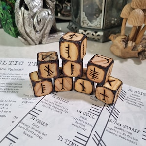 May include: Six wooden dice with Celtic Ogham symbols burned into the surface. The dice are arranged in a pyramid shape on a white surface with a chart of Ogham symbols and their meanings.