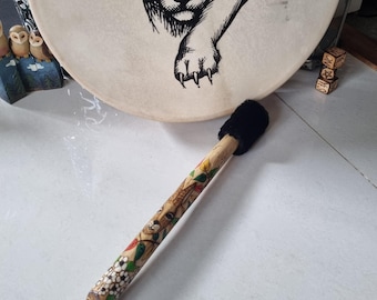 May include: A round frame drum with a cream-colored head featuring a black ink illustration of a mountain lion. A wooden mallet with a black fuzzy head and a hand-painted handle decorated with floral motifs and a small face rests in front of the drum.