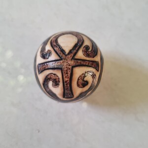 May include: A wooden bead with a brown ankh symbol carved into it. The bead is round and has a light brown colour.