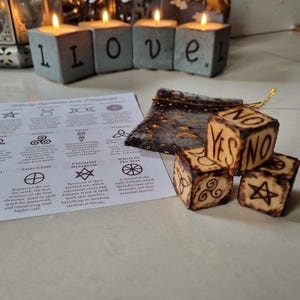 May include: Four wooden dice with symbols and the words "YES" and "NO" burned into the surface. The dice are sitting on a white surface with a piece of paper with symbols and text about witchcraft.