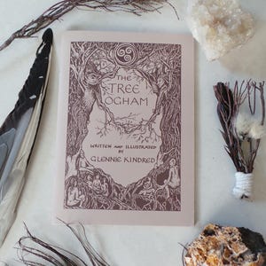 May include: A softcover book titled The Tree Ogham written and illustrated by Glennie Kindred. The cover features a brown ink illustration of trees and figures on a light tan background with a triskelion symbol at the top centre.