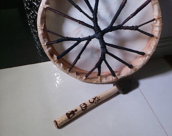 May include: A brown wooden frame drum with a white leather head and black leather lacing. The drum is being played with a brown wooden beater with a brown felt head. The beater has a symbol of Om and a crown carved into it.