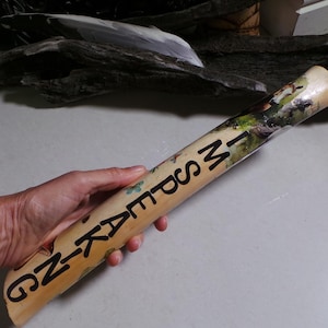 May include: A cylindrical wooden object with the word "KAYAKING" printed in black. The object has a painted landscape scene and is held in a hand. The background includes driftwood.