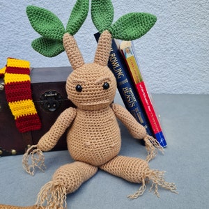 Könnte beinhalten: Eine gehäkelte Alraune, eine fiktive Pflanze aus der Harry-Potter-Reihe, mit einem beigefarbenen Körper, grünen Blättern und beigen Wurzeln. Die Alraune befindet sich vor einer Truhe, einem Schal und Büchern.