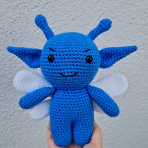 Könnte beinhalten: Eine blaue gehäkelte Fee mit weißen Flügeln. Die Fee hat einen schelmischen Ausdruck mit schwarzen Augen und einem kleinen Lächeln.
