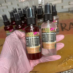 May include: Two small glass bottles of facial toner with spray tops. The bottle on the left is red and labelled "Aloe Rose Facial Toner". The bottle on the right is clear and labelled "Cucumber Mint Facial Toner".