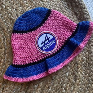 Puede incluir: Sombrero de cubo de crochet rosa, azul y negro con un parche redondo azul y blanco que dice "Alpine" y tiene el número "4".