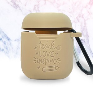 May include: A tan silicone case for AirPods with a carabiner clip. The case has the words "teach love inspire" and a pencil and apple graphic.