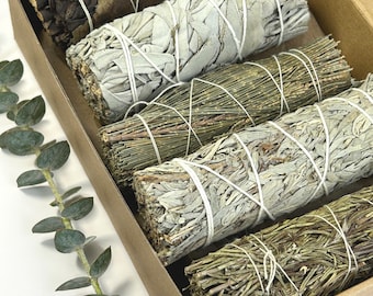 May include: A collection of sage bundles in a cardboard box. The bundles are wrapped with white string and vary in color from light green to brown. A sprig of eucalyptus is in the lower left corner.