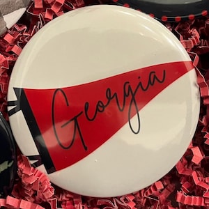 May include: A white button with a red pennant design featuring the word "Georgia" in a cursive font. The button is surrounded by red paper shreds.