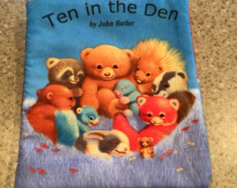 May include: A blue book titled "Ten in the Den" by John Butler. The cover features a group of cuddly toys, including a bear, a raccoon, a hedgehog, a badger, a rabbit, and a fox.