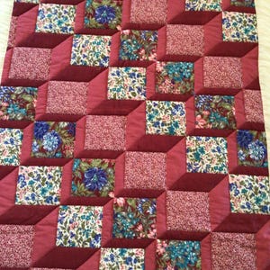 Op de afbeelding: Een quilt met een 3D-effect, met een patroon van vierkanten met bloemenmotieven in tinten roze, blauw en groen. De vierkanten zijn geplaatst op een bordeauxrode achtergrond.