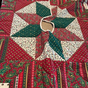 May include: A circular quilted tree skirt featuring a patchwork design of red, green, and cream fabrics. The pattern includes star shapes, paisley prints, and floral motifs with a central opening and a slit for placement around a tree base.