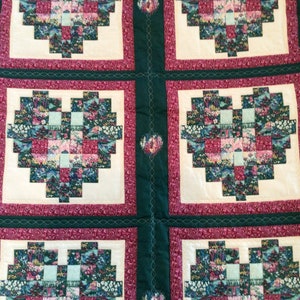 Op de afbeelding: Een quilt met een groene achtergrond en zes witte vierkanten met rode randen. Elk vierkant bevat een patchwork hartontwerp in tinten groen, blauw, roze en wit.