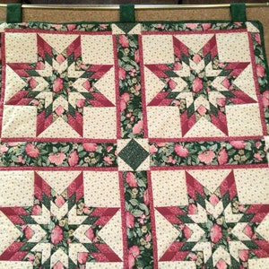 以下が含まれることがあります： ピンク、グリーン、ホワイトの星形模様のキルティングされた壁掛け。生地は花柄です。