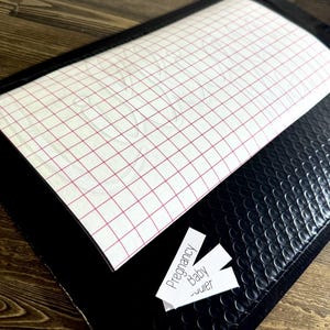 May include: A black bubble envelope with a white sheet of transfer paper featuring a red grid pattern. Three small white labels read "Pregnancy", "Baby", and "...ier". The envelope is on a wooden surface.