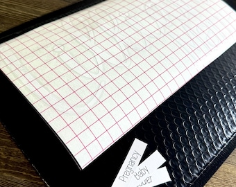 May include: A black bubble mailer with a white sheet of transfer paper with a red grid pattern. Three small white labels read "Pregnancy", "Baby", and "...ier". The mailer is on a wooden surface.