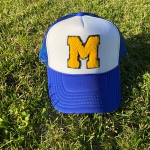 May include: A blue and white trucker hat with a yellow "M" patch. The hat has a blue brim and mesh back, with a white front panel. The letter "M" is in a gold, textured material.