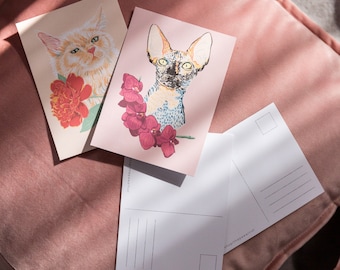 May include: Three postcards featuring images of cats. The first postcard shows a white cat with orange markings and a red flower. The second postcard depicts a hairless cat against a pink background with pink flowers. The third postcard is blank with lines for writing.