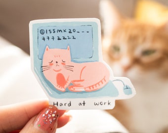 Könnte beinhalten: Ein Aufkleber mit einer rosa Katze, die auf einem blauen Laptop schläft, mit dem Text "Hard at work" und dem Code "O155MX20---77772222" auf dem Bildschirm.