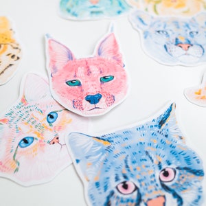 May include: A set of colourful cat head stickers with different patterns and colours. The stickers feature a variety of cat breeds, including a tabby, a calico, and a Siamese.