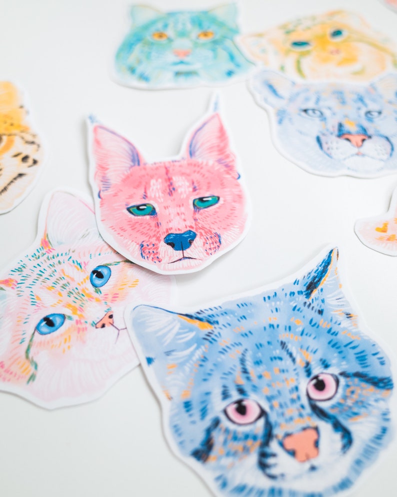 May include: A set of colourful cat head stickers with different patterns and colours. The stickers feature a variety of cat breeds, including a tabby, a calico, and a Siamese.