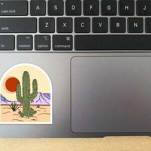 Pu&ograve; includere: Un adesivo raffigurante un paesaggio desertico con un grande cactus verde, un sole rosso e montagne viola. L'adesivo ha la forma di un arco bianco, con uno sfondo beige. L'adesivo &egrave; posto su un laptop.