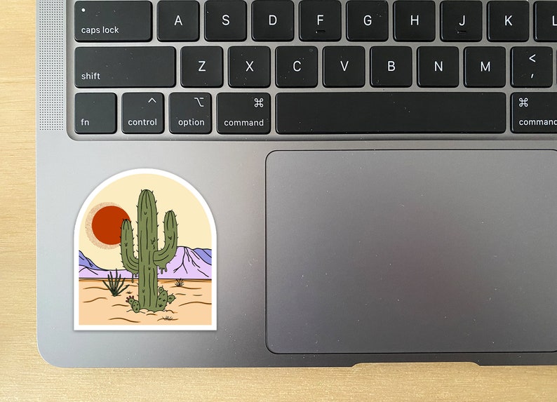 Pu&ograve; includere: Un adesivo raffigurante un paesaggio desertico con un grande cactus verde, un sole rosso e montagne viola. L'adesivo ha la forma di un arco bianco, con uno sfondo beige. L'adesivo &egrave; posto su un laptop.