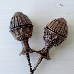 Könnte beinhalten: Zwei antike, eichelförmige Möbelknöpfe aus Bronze mit dekorativen Details. Jeder Möbelknopf hat eine Schraube zur Befestigung. Die Möbelknöpfe sind warmbraun und liegen auf einer weißen Oberfläche.