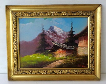 Könnte beinhalten: Ölgemälde einer Berglandschaft mit einem Haus, gerahmt in einem goldenen, verzierten Rahmen. Das Gemälde zeigt eine Bergkette mit schneebedeckten Gipfeln, ein Haus und einen Weg durch ein grünes Feld. Der Himmel ist blau mit weißen Wolken.